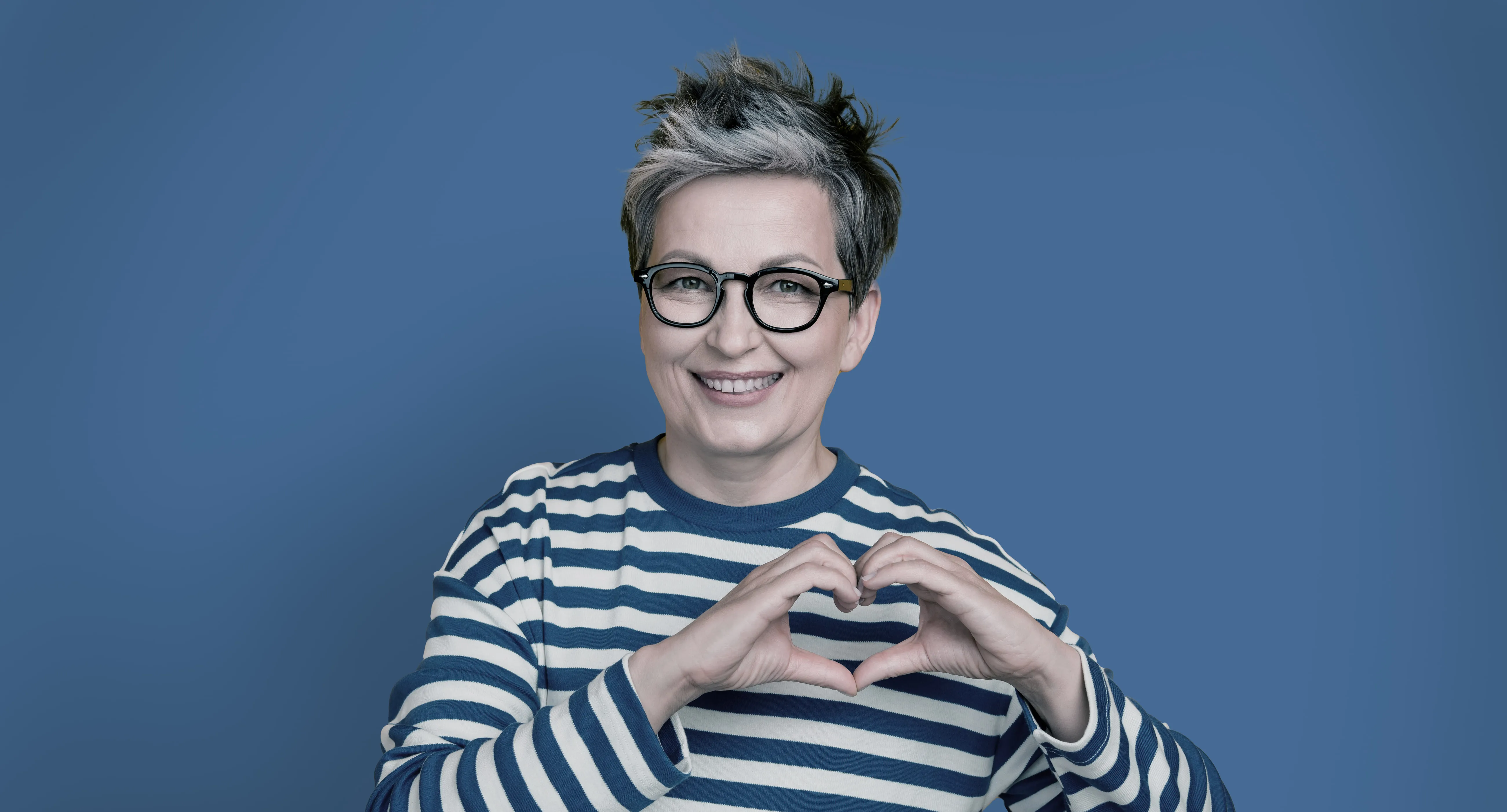A smiling client in glasses and a striped top making a heart with her hands