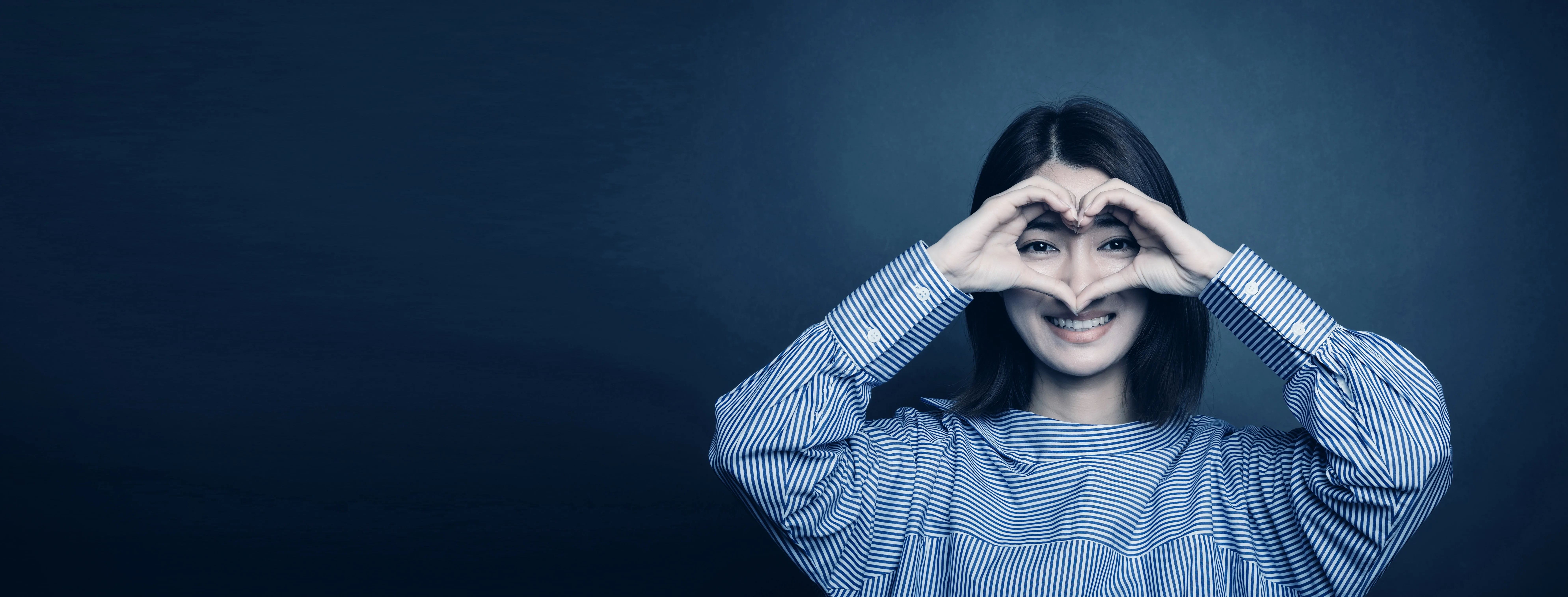 A client making the heart-hands gesture around her eyes, smiling, against a deep navy studio backdrop
